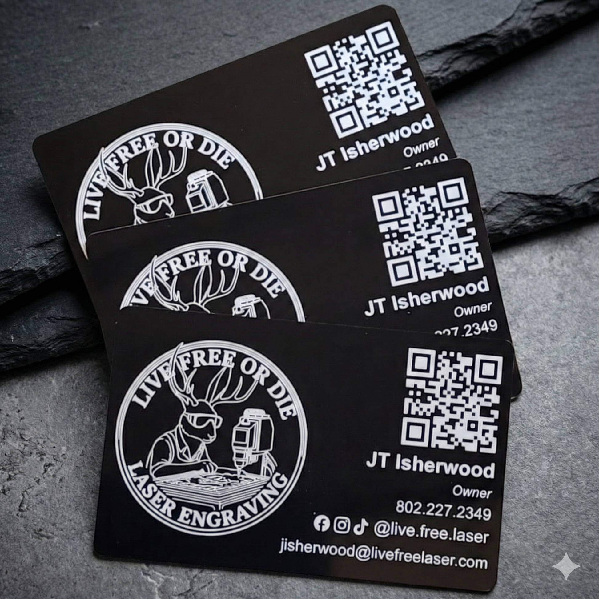 Close-up of three custom laser-engraved black metal business cards fanned out on a dark stone background. Demonstrates high-contrast bright silver text, a complex logo, and a scan-ready QR code etched directly into the anodized aluminum.